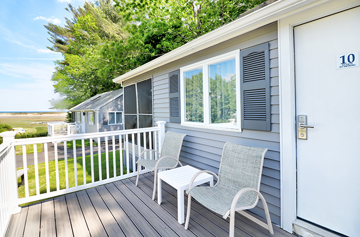 A porch with two chairs and a small table on a deck with a white railing, in front of a building with gray siding and a white door.