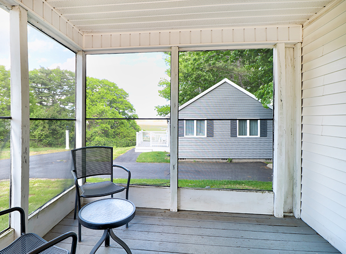A screened-in porch with a small table and chair, looking out onto a street and neighboring house.
