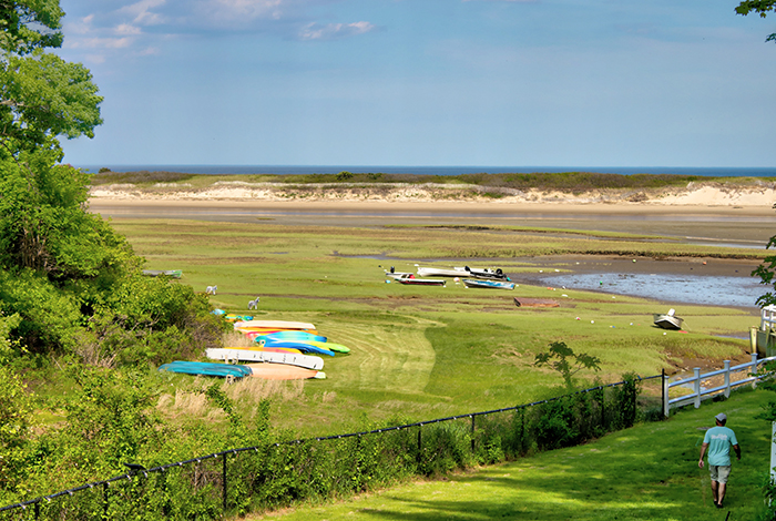 A view of a beach with boats on the grass and a person walking away in the distance.