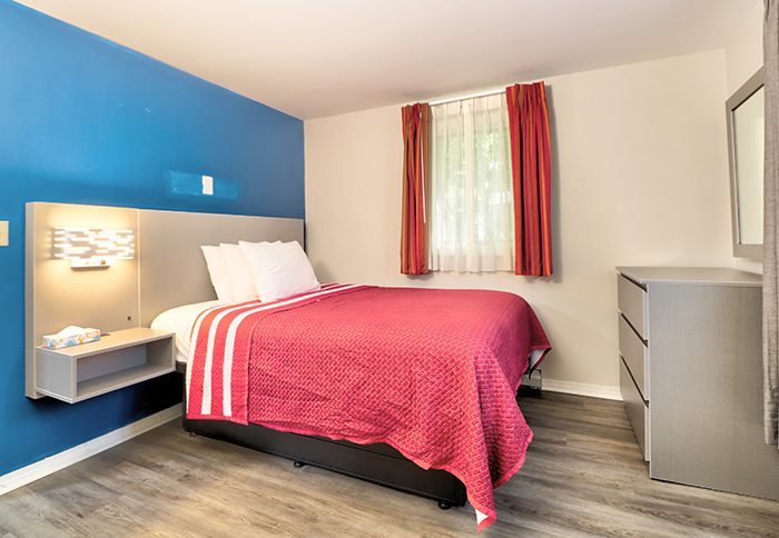 A hotel room with a red bedspread, white pillows, a gray dresser, and a window with red curtains.